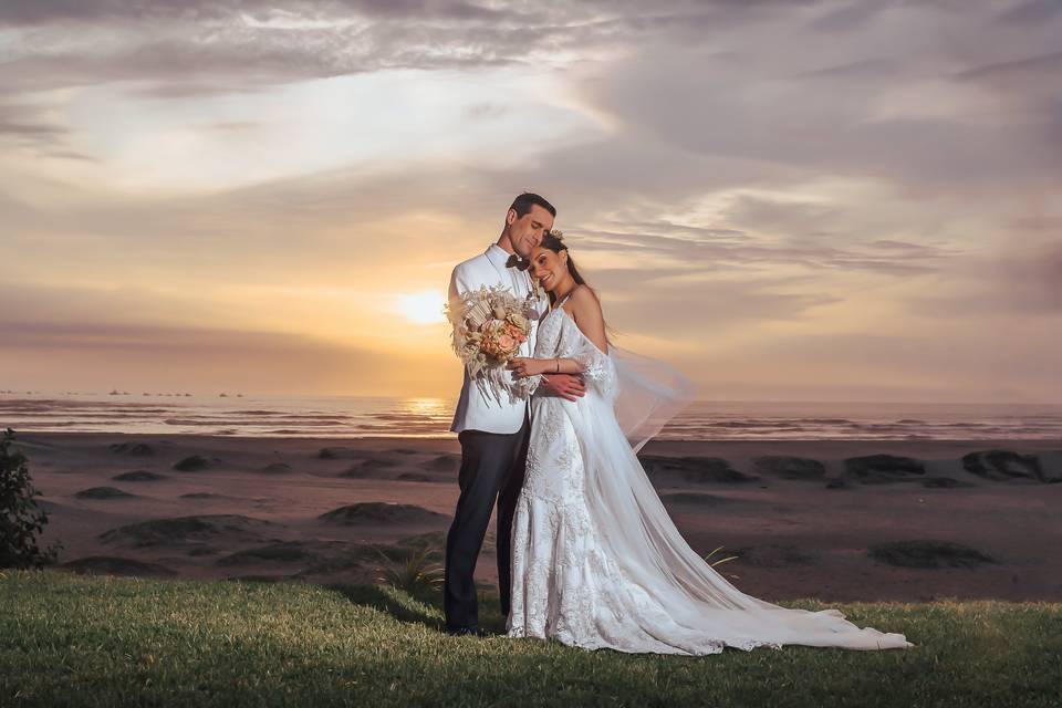 novios abrazados frente a la playa en pleno atardecer la novia sujeta su bouquet de novia