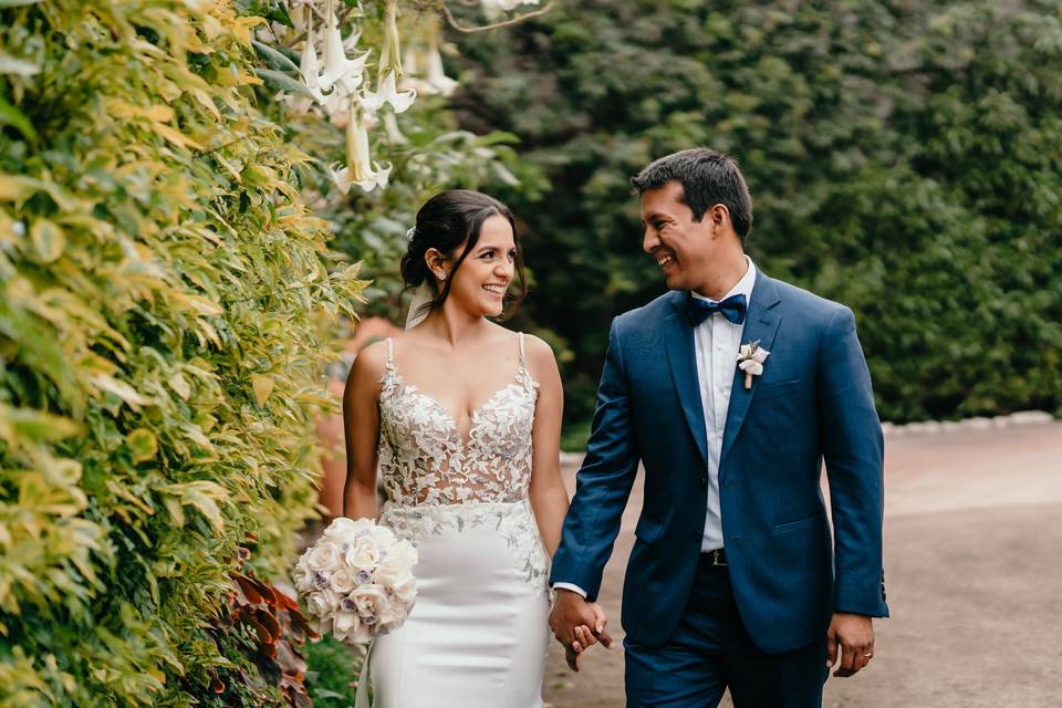 novio con terno azul y novia con bouquet blanco sonriendo tomados de la mano mientras caminan en medio de árboles y flores