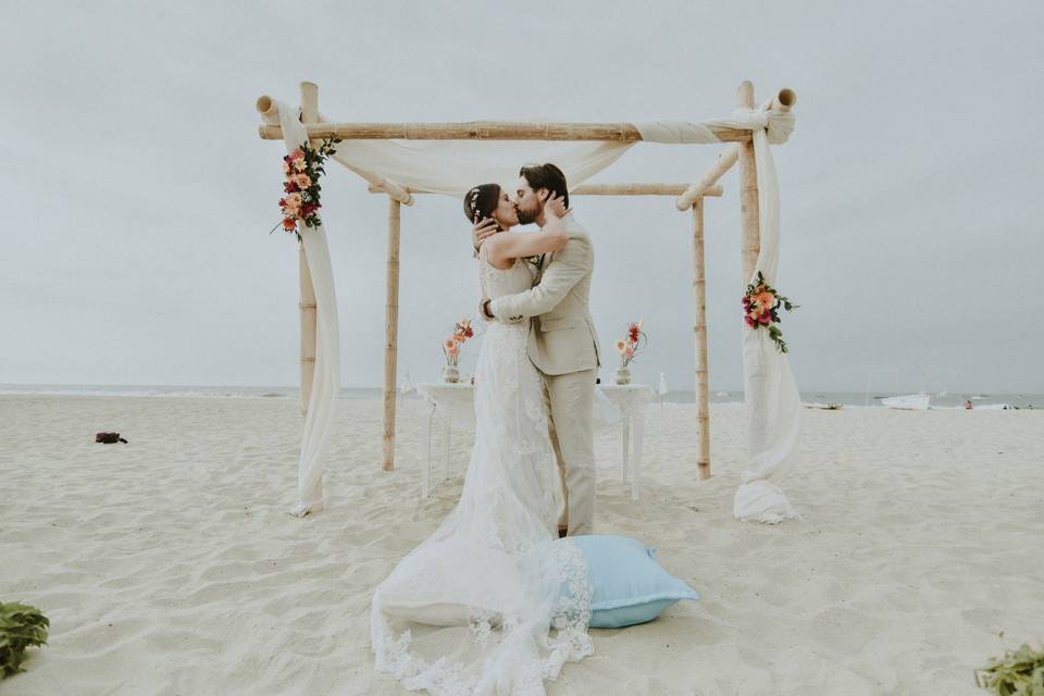 chico y chica vestida de novia besándose delante de un altar de madera, flores y tul en la playa