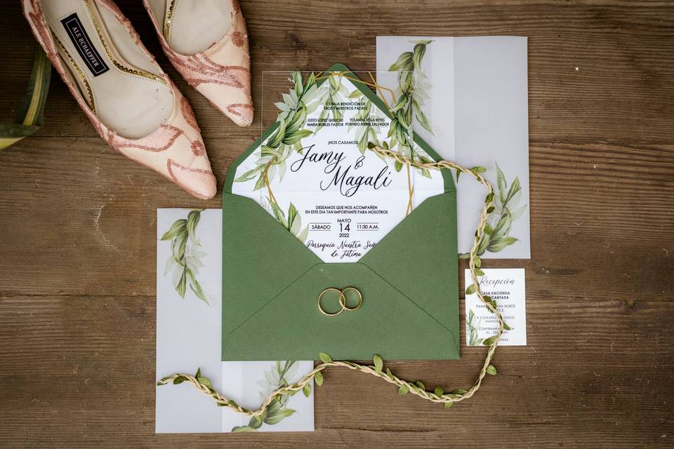 tarjeta de matrimonio acrílica con motivos de hojas verdes con sobre verde olivo al lado de zapatos de novia