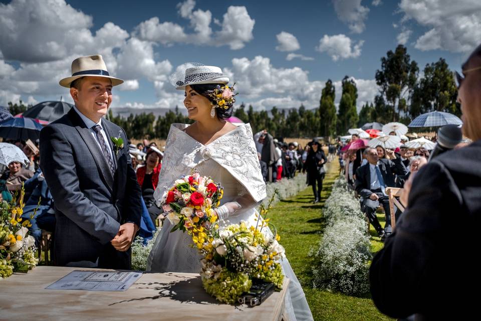 chico con terno azul y sombrero junto a chica con vestido de novia y flores delante del altar al aire libre