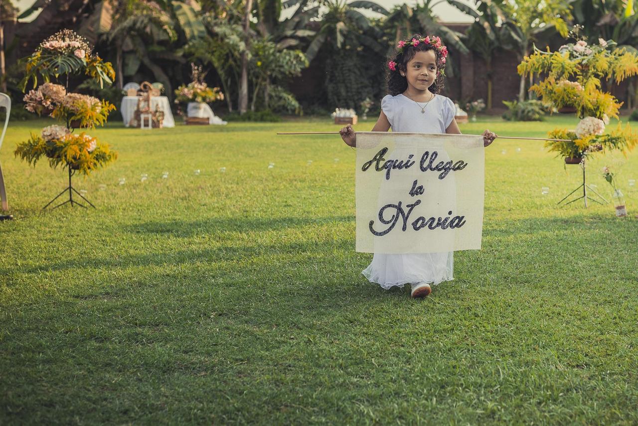 niña con corona de flores llevando un letrero que dice aquí llega la novia